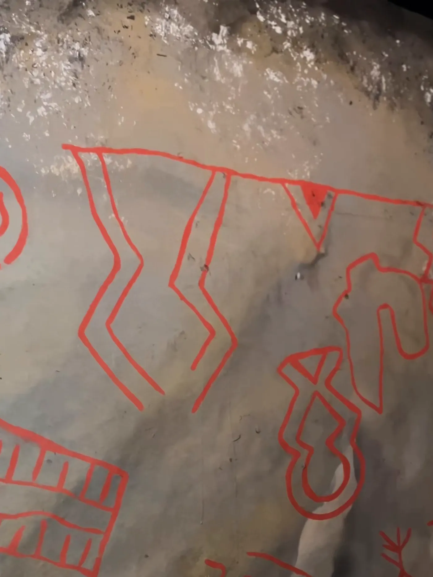 Parede de pedra com pinturas rupestres em vermelho: figuras geométricas e humanas.